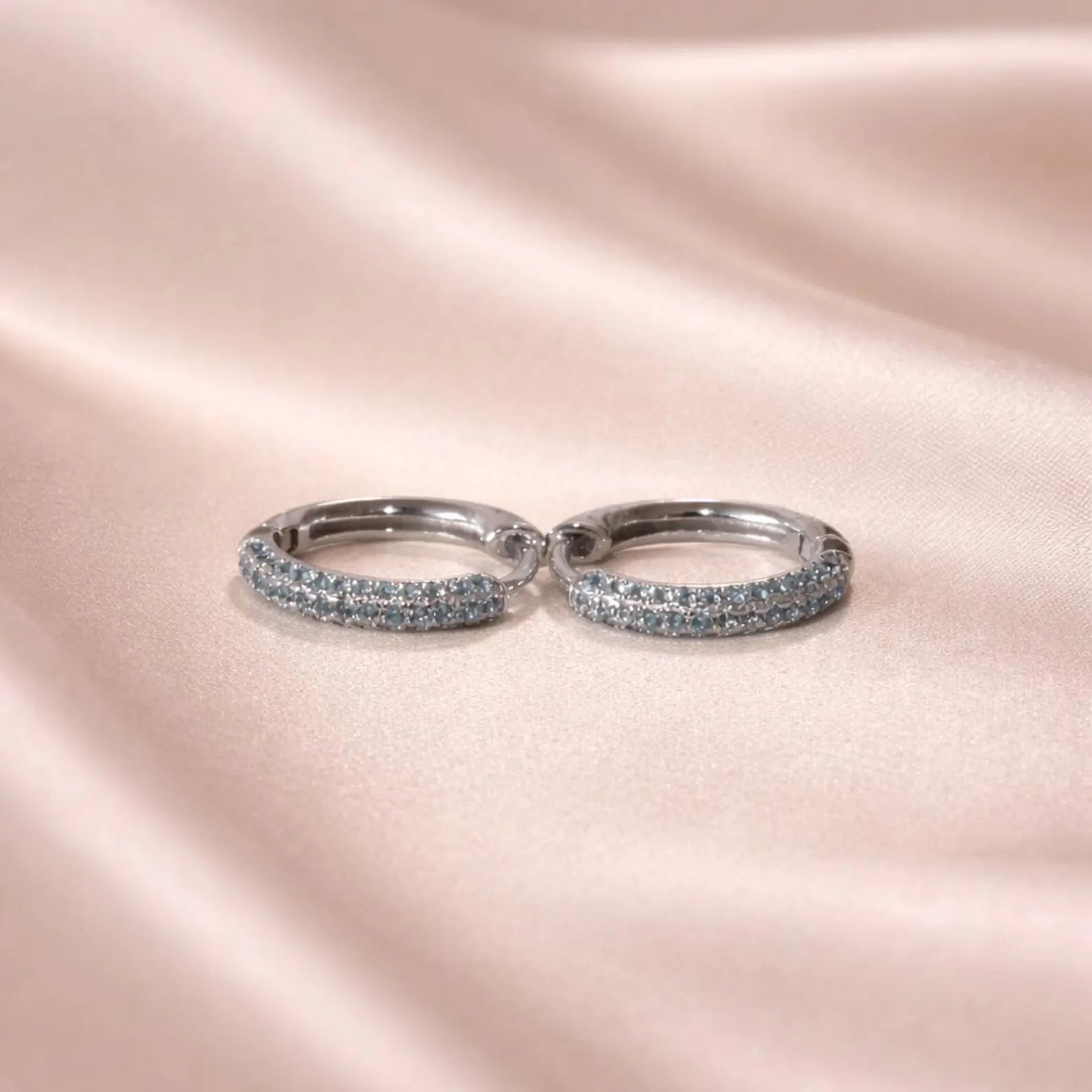 Pair of silver aquamarine hoop earrings with embedded stones on a pink satin background